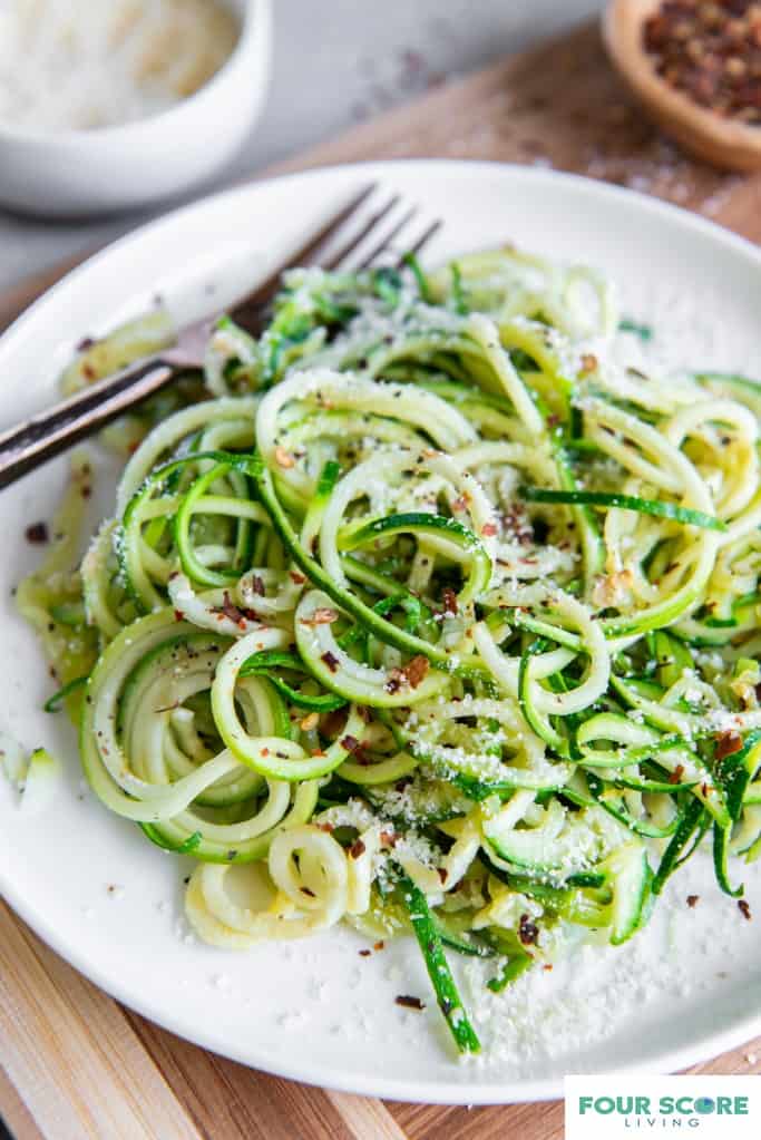 Diagonal aerial view of spiralized, cooked zucchini noodles with Parmesan cheese and red pepper flakes on a white plate resting on a wooden cutting board with a dish of parmesan cheese and a small wooden dish of red pepper flakes in soft focus in the background.  