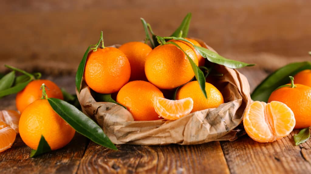 On a wooden surface, whole clementine with skin on, some with green leaf, and some peeled clementine, stacked in a flat brown paper bag with 6 clementine out of the bag. wood background.