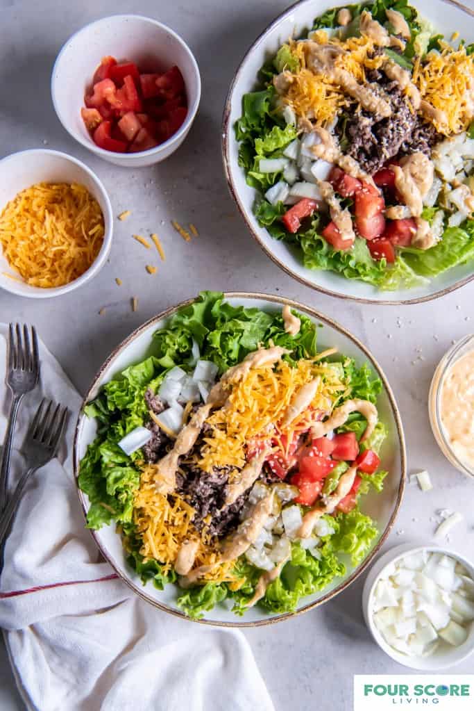 Two plates of salad greens with, each plate containing cooked ground beef, shredded cheddar cheese, diced red tomatoes, diced white onions, drizzled 1000 island sauce and a sprinkle of sesame seeds. In the background there is a small white dish with chopped onion, a glass bowl with 1000 island dressing, a white dish of shredded cheddar cheese and a white dish of diced red tomatoes. Two forks and a white dish towel, all placed on a white background.