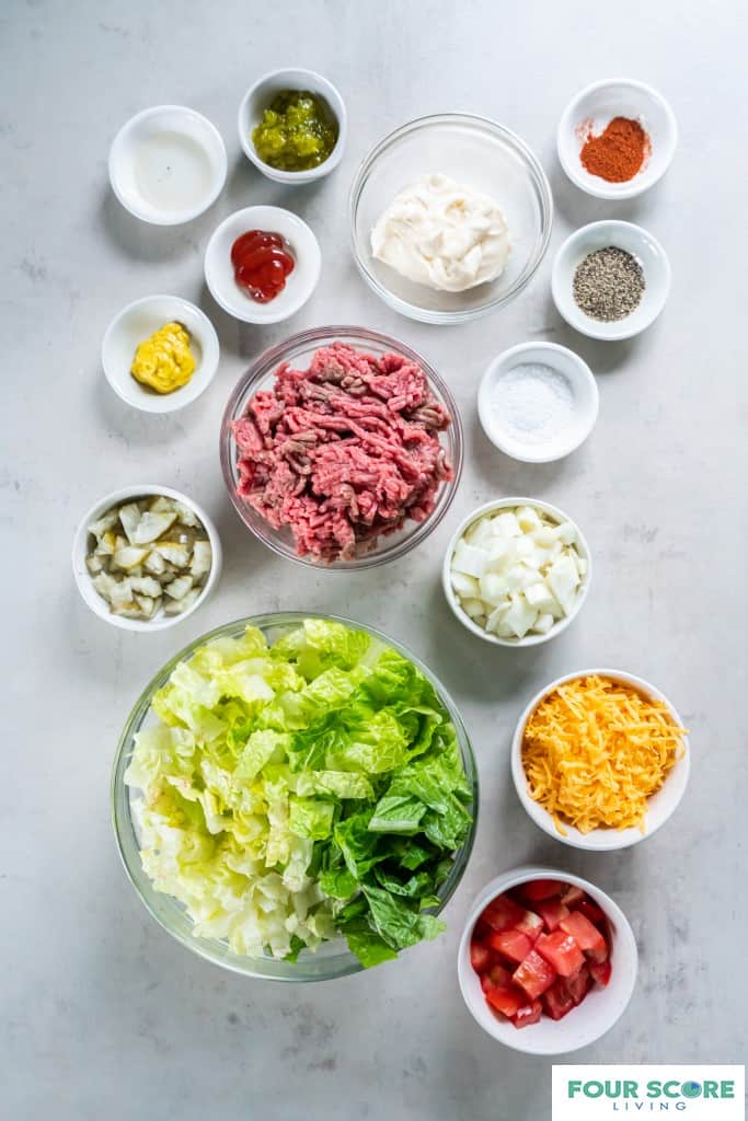 Glass bowls and small white bowls containing ingredients to create a big mac salad, including, diced tomatoes, chopped lettuce, shredded cheddar cheese, chopped white onion, raw ground beef, salt, pepper, mustard, ketchup, pickled relish, white vinegar and paprika. 