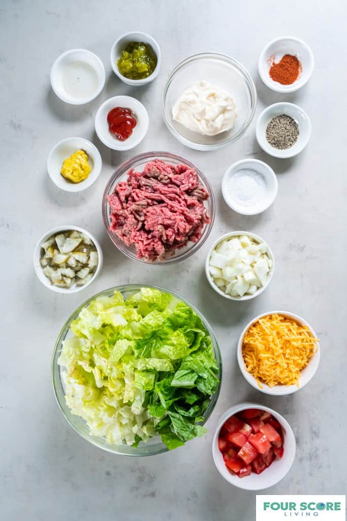 Glass bowls and small white bowls containing ingredients to create a big mac salad, including, diced tomatoes, chopped lettuce, shredded cheddar cheese, chopped white onion, raw ground beef, salt, pepper, mustard, ketchup, pickled relish, white vinegar and paprika.