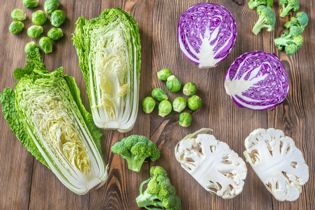 A head of romaine lettuce sliced in half with placed on a brown in color background with wood graining, with inside of the romaine facing up. Two clusters of about 10 whole Brussel sprouts, one whole white cauliflower cut in half with the insides facing up and 7 florets of broccoli all on a brown tone wood grain surface.  