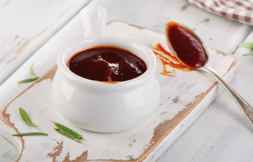 BBQ sauce in a white bowl, placed onto a white cutting board layered on a white wooden surface, with a spoon full of BBQ sauce, fresh green garnish and a brown checkered towel in the background.