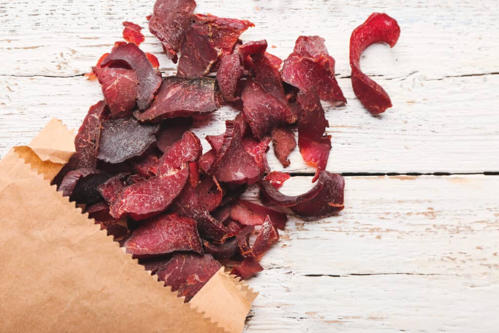 Brown paper bag with cut strips of beef jerky spilling out onto a rustic wooden, whitewashed surface.