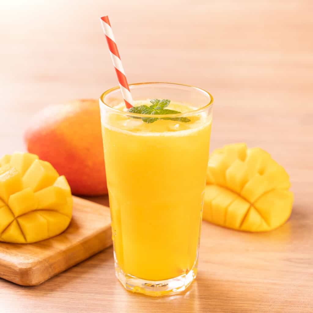 Tall glass of bright yellow mango juice with four fresh mint leaves as a garnish and a white and red straw with one whole unpeeled mango in the background and 2 halves of a fresh mango with the flesh diced and the flesh side turned out, one half placed on a wooden cutting board and all resting on a light toned wood surface.
