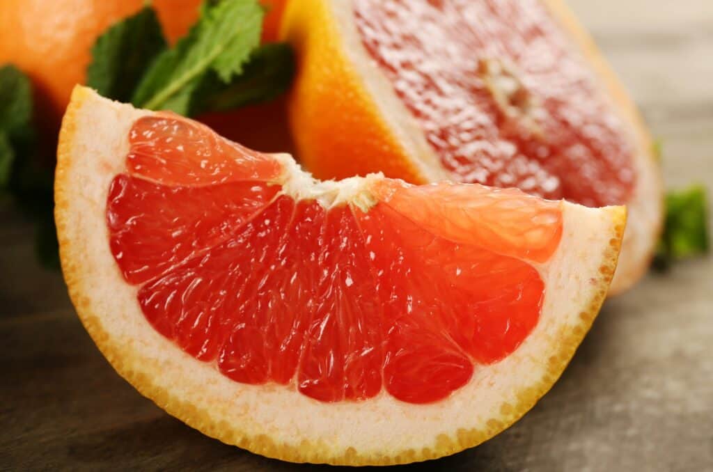 A sliced wedge of pink grapefruit, with a half of a grapefruit and a whole unpeeled grapefruit as well as leafy green garnish in the background, all placed on a rustic wood surface. 