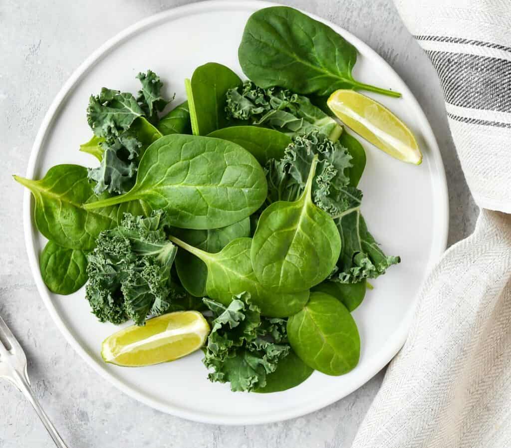 White plate of fresh spinach and curly kale with 2 slices of line with a silver fork and a white dish towel with a grey stripe in the background, all placed on a white stone surface.