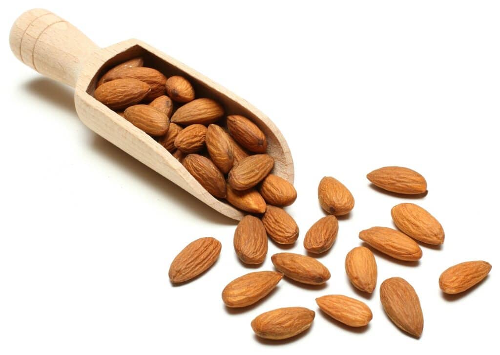 Front diagonal view of a small natural wooden scoop overflowing with whole almonds on a white background.