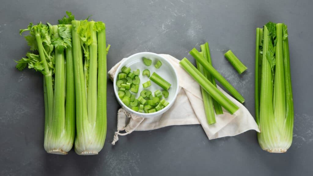 Aerial view of two heads of fresh green celery on the left with a small white bowl of ½ inch chopped celery pieces inside and four celery ribs placed outside the bowl on top of a natural colored cloth bag. One head of fresh celery is on the right . All on a dark grey stone background.
