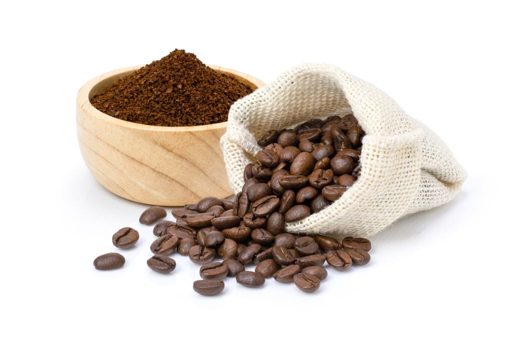 Small natural wood light in color bowl filled with fresh coffee grounds with a natural small sac filled with whole coffee beans spilling out onto the surface, all on a white background.