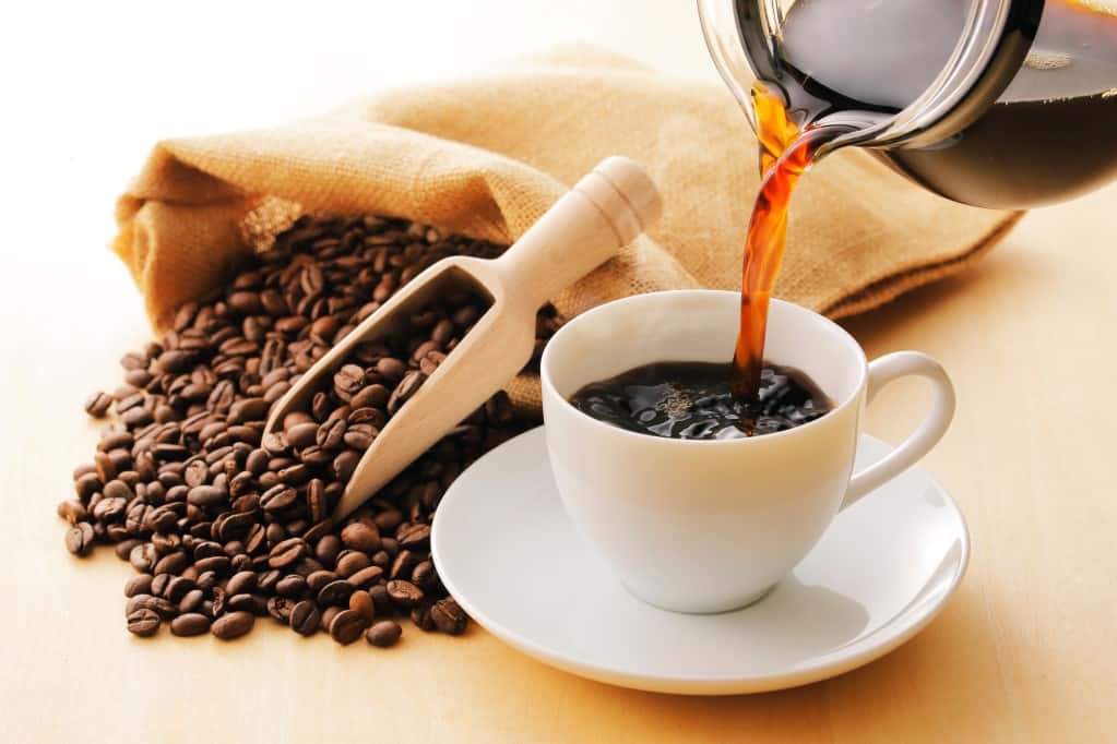 White cup and saucer with a glass coffee pot pouring steaming coffee into the cu with a natural sac of whole coffee beans overflowing onto a wooden surface and a natural wood scoop. 