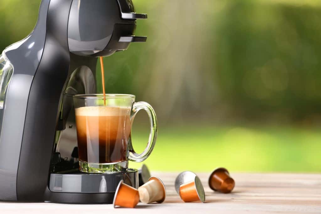 Coffee machine brewing coffee from a pod into a clear glass mug with a handle with 6 coffee pods spilled out on a wooden surface with a soft focus green nature scene in the background.