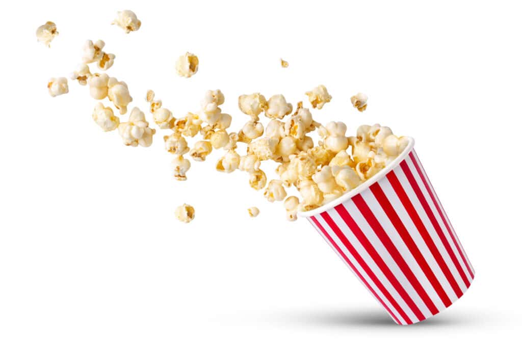 Red and white vertical striped round tub of popcorn tipped to the right side with popped popcorn spilling out of the top and heading upwards in midair on a white background.