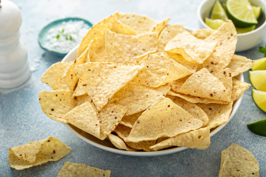 Medium sized white bowl overflowing with tortilla corn chips with fresh lime wedges on the surface and a small dish of lime wedges in the right background. In the left background is a small blue dish of corn starch in soft focus and a white wooden pepper mill grinder.