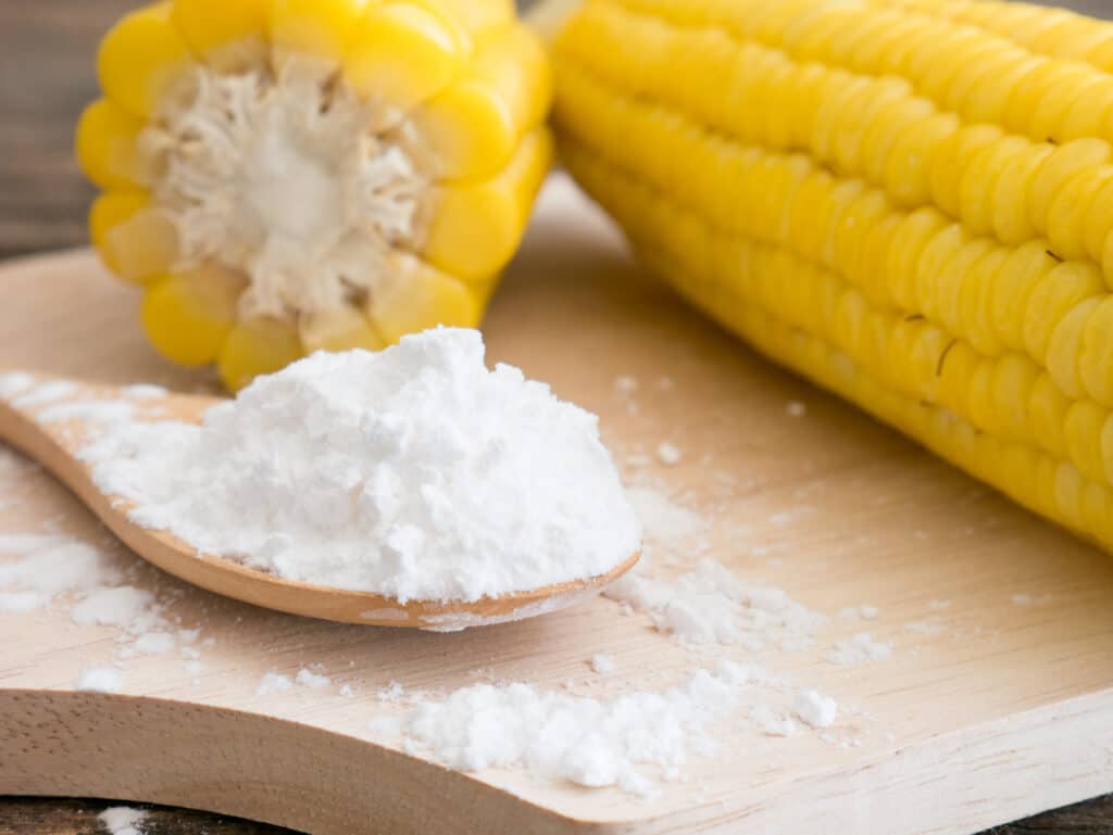 Close up frontal view of a light colored wooden spoon filled with corn starch that is overflowing onto the light wooden cutting board surface with two yellow corn on the cobs in soft focus in the background.