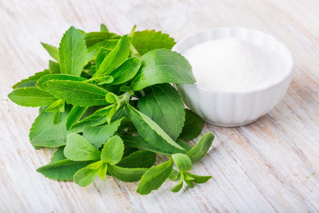 Small white bowl of white finely granulated stevia sweetener placed next to a handful of fresh green stevia leaves all resting on a rustic, whitewashed surface.