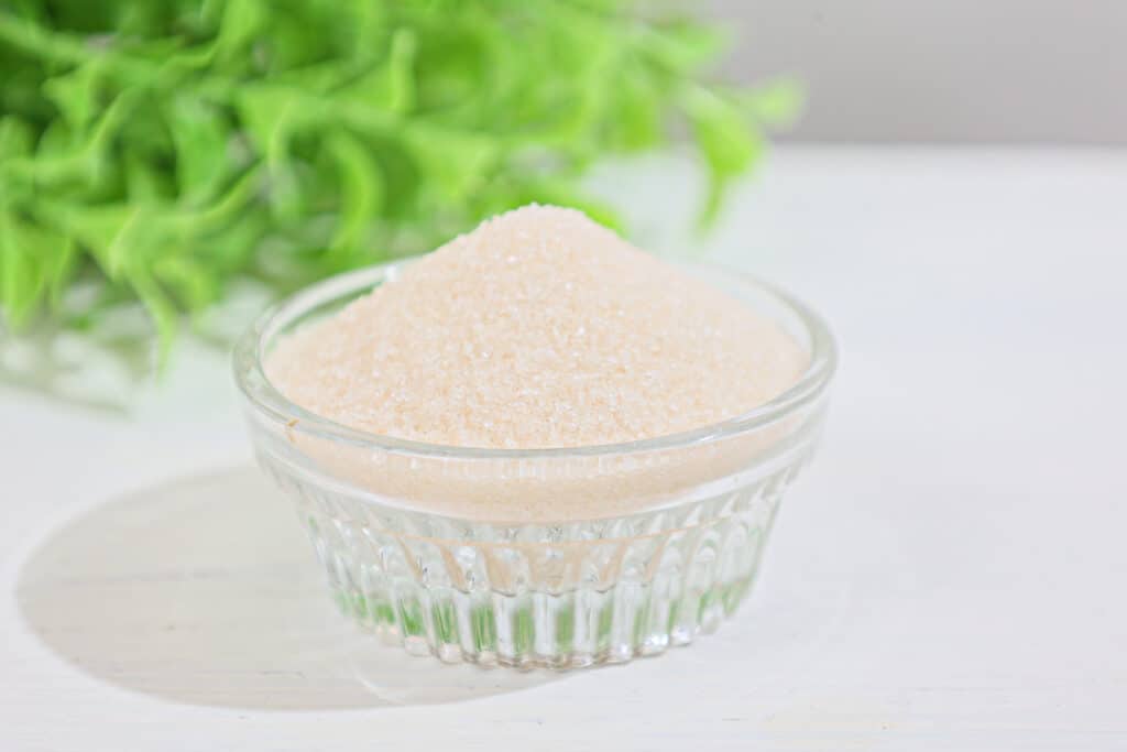 Granulated monk fruit sweetener in a clear glass dish with fresh green leaves in the background with a soft focus, all on a white white washed surface.