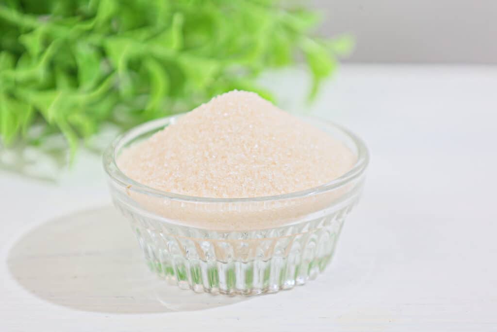 Small clear glass round dish filled with ivory colored xylitol crystals with a large bunch of fresh xylitol leaves in soft focus in the background, all placed on a white surface.