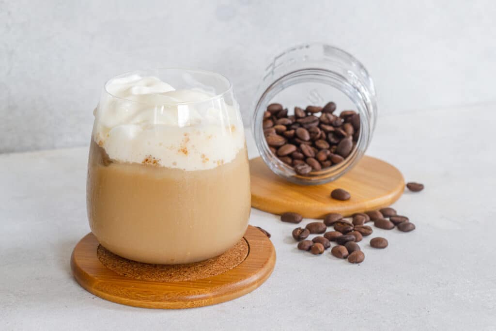 Small glass of expresso and milk topped with whipped heavy cream  placed on a medium colored wood and cork coaster. A small glass jar of whole expresso beans tipped on its side spilling beans onto the surface, with jar placed on a round wooden disc, all on a white stone background. 