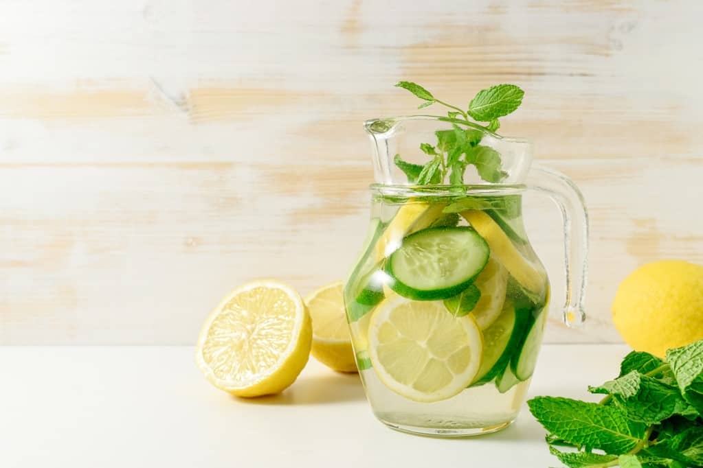 Clear glass pitcher of water filled with sliced lemon, cucumber, sprigs of mint and two lemons on the surface one sliced in half, one whole, and a medium bunch of mint leaves on an ivory surface with a light wooden white washed background.