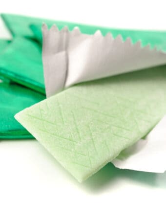 Stick of pale green chewing gum and the green wrapping foil on top of a pile of unwrapped sticks of chewing gum in green foil on a white background.