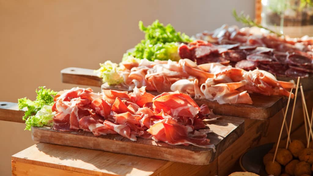 Four rectangular cutting boards placed along a wood board banquet style, each cutting board is piled with different types of sliced ham and a green leafy garnish.
