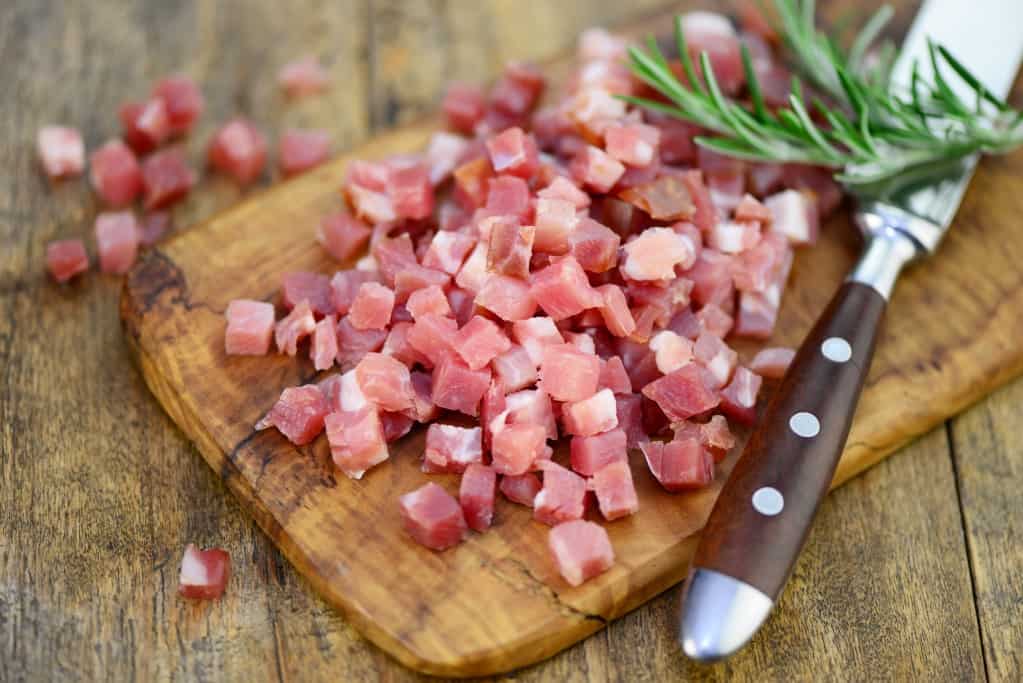 Diced thick cut of ham piled on a medium brown cutting board with a large steel knife and a large spring of rosemary, with the diced ham spilling onto the rustic wood background surface.