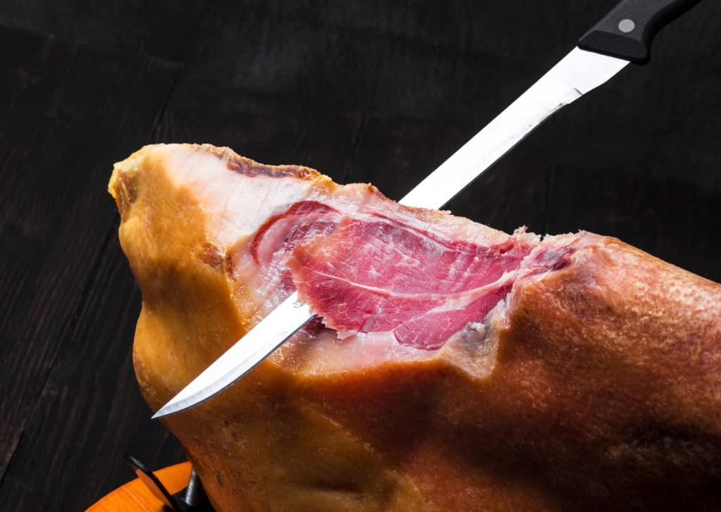 Diagonal view of a long steel knife slicing traditional Spanish ham from the leg with a dark background.