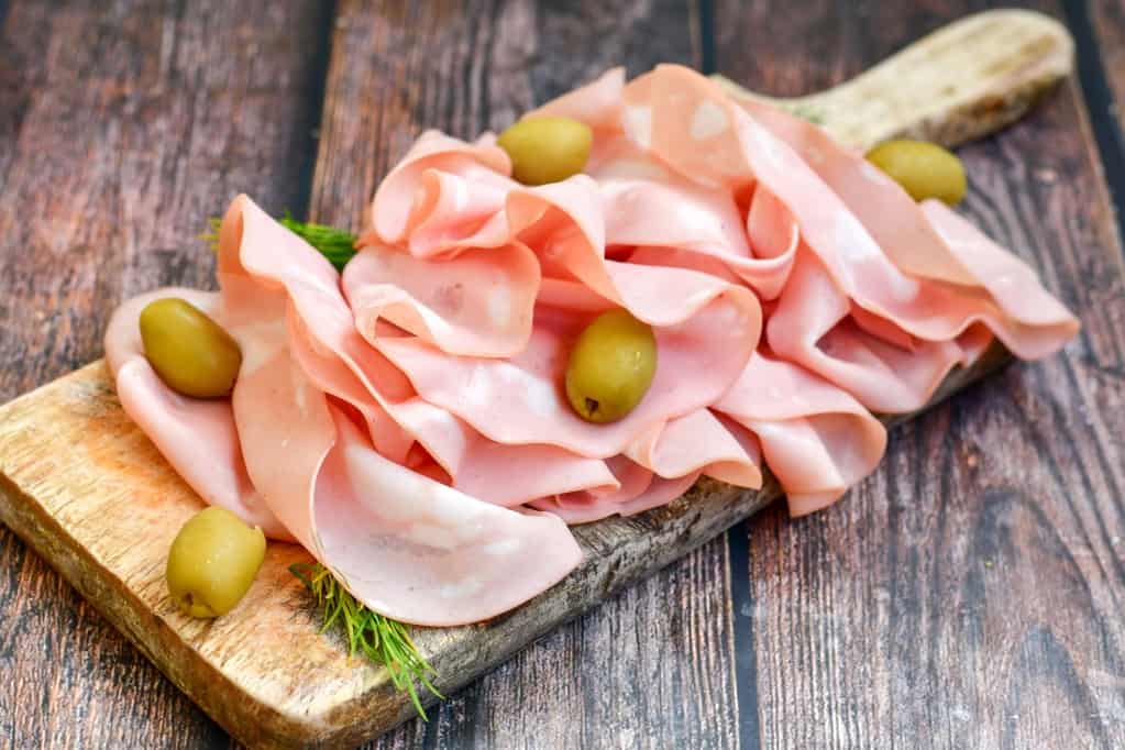 Rectangular cutting board with a handle piled with sliced ham and garnished with whole green olives and sprigs of fresh herbs. 