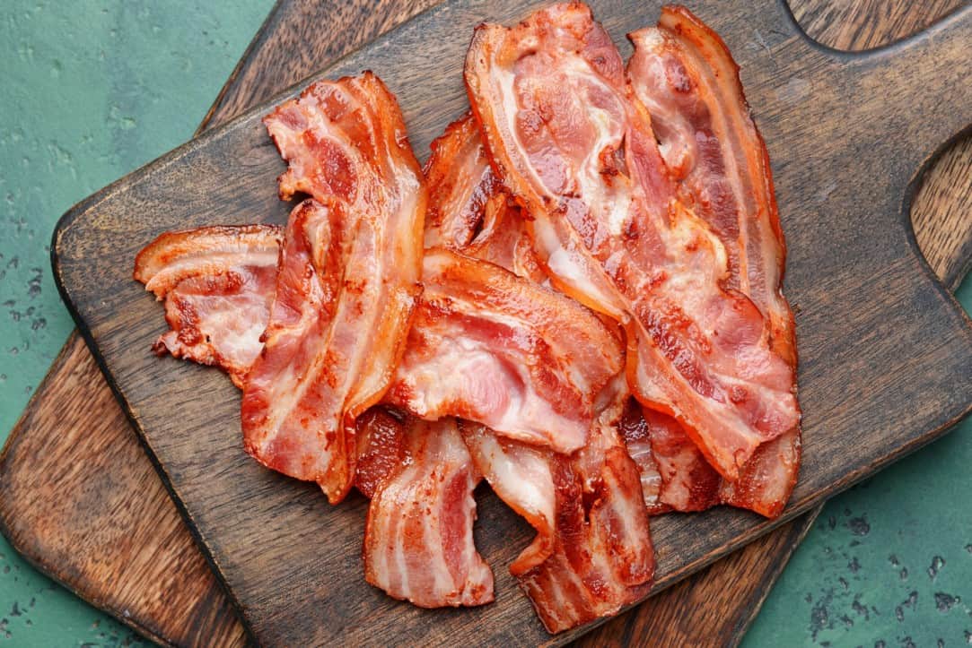 Approximately 10 strips of fried bacon placed on top of two stacked wooden cutting boards sitting on top of a rustic painted surface.