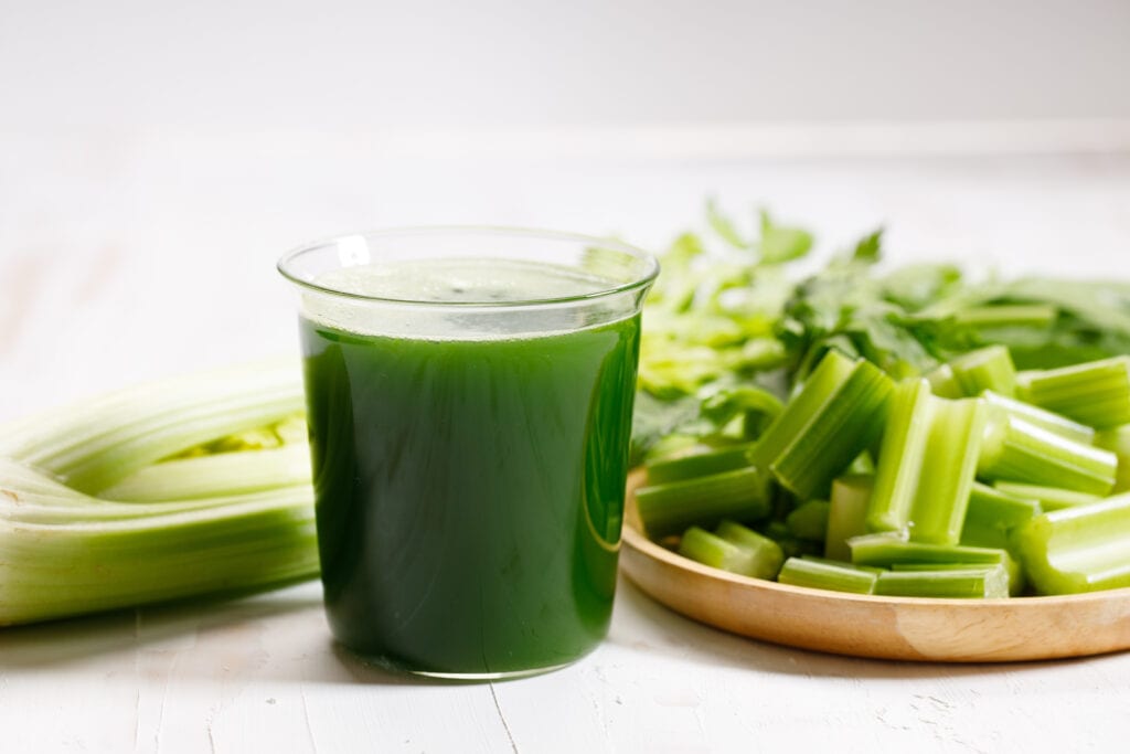 Short clear glass of green celery juice in the center with a light colored wooden plate of celery chopped into 2-inch segments on the right and a head of celery on the left extending behind the glass of juice with the celery leaves in soft focus in the background.