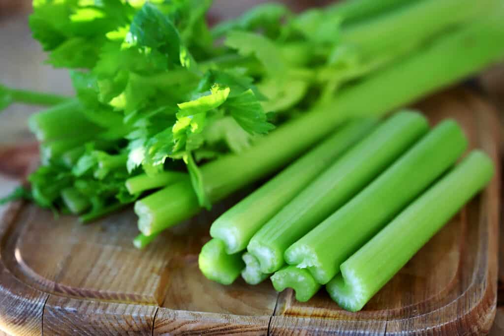 Celery ribs or stalks on a wooden board with fresh celery leaves in soft focus in the background.