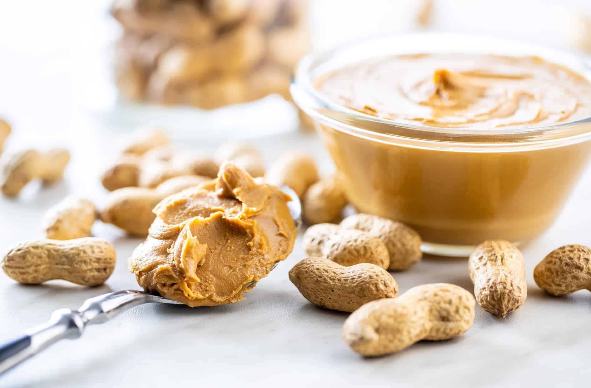 Front closeup view of a clear glass dish filled with peanut butter, a spoon nearby filled with peanut butter, peanuts in the shell scattered on the surface and a jar of peanuts in the shell in soft focus in the background, all resting on a light colored stone surface.
