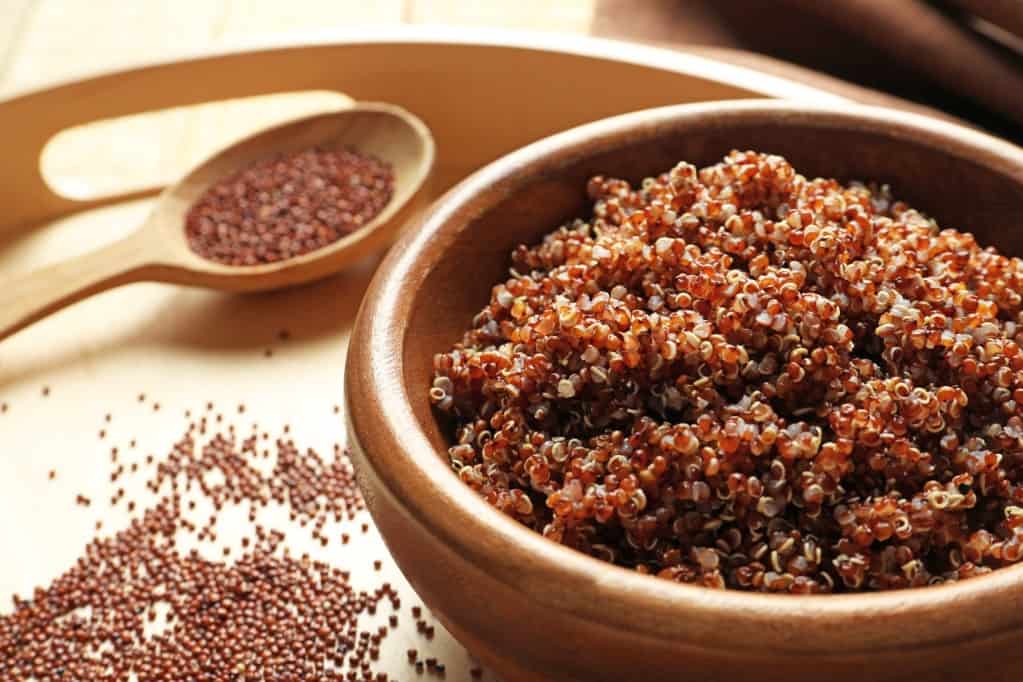 Wooden bowl with boiled red quinoa grains  on a light wooden tray with uncooked red quinoa spilled on the tray and a medium sized light wooden spoon in soft focus partially filled with uncooked red quinoa. 