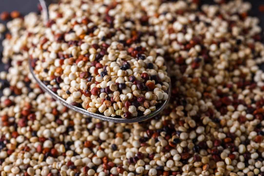 Up close diagonal shot of mix of uncooked quinoa grains spilled out on the surface of a dark stone background filling the frame, with a medium wooden spoon overflowing with Quinoa in the foreground.