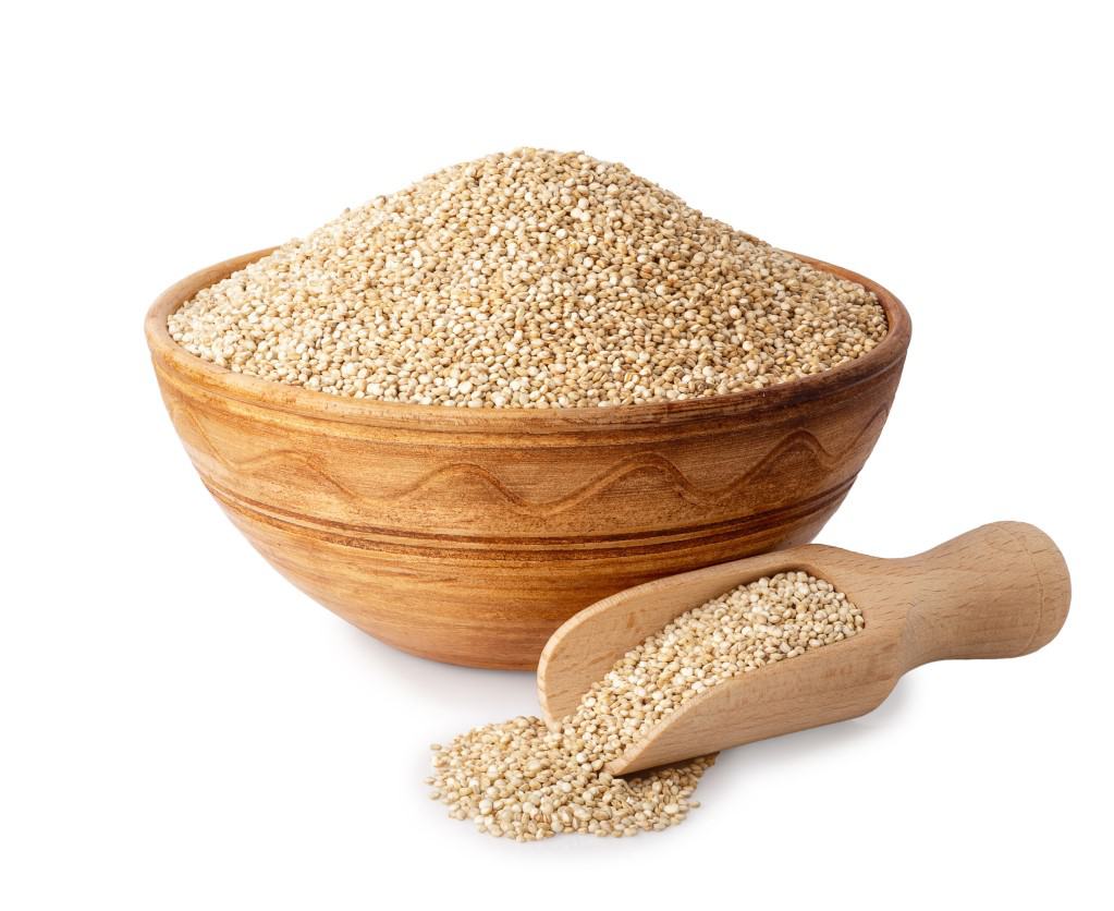 White uncooked quinoa grains in a light wood tone ceramic bowl and natural wooden scoop with white uncooked quinoa spilling out on an isolated on white background.