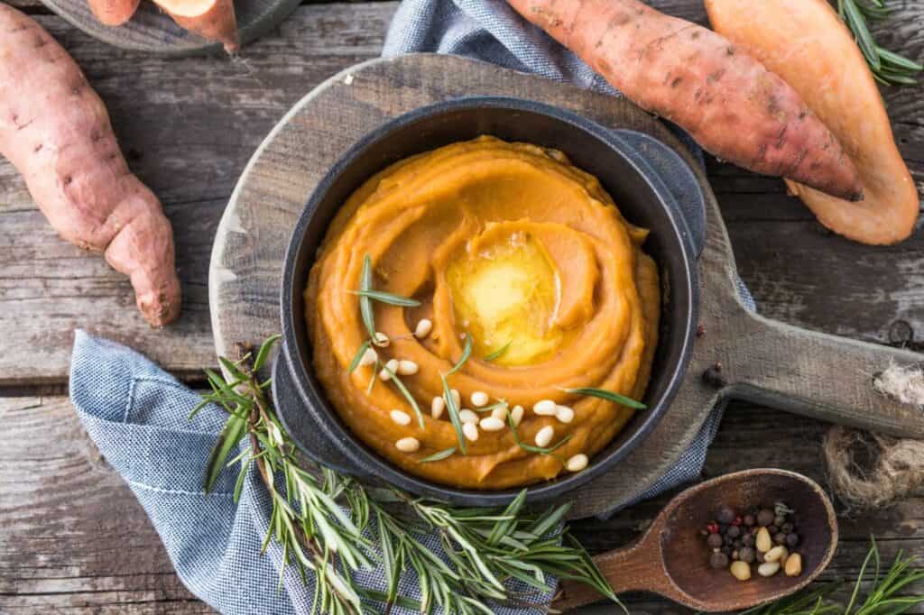 Aerial view of mashed sweet potato or sweet potato puree with rosemary in a an iron pan placed on a round serving board with a handle set on a rustic wood table surface. A denim blue napkin, springs of fresh rosemary, a medium wooden spoon with whole spice kernels and whole raw sweet potatoes, including one half of a raw sweet potato sliced lengthwise.