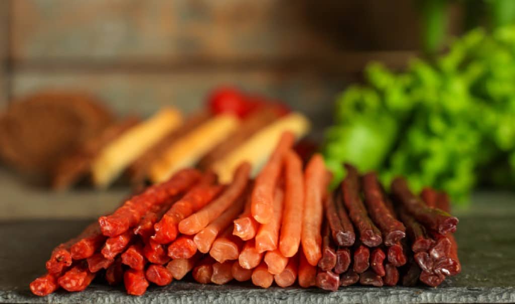 There colors of stick jerky piled onto of a slate surface with green and yellow vegetables in the background. 