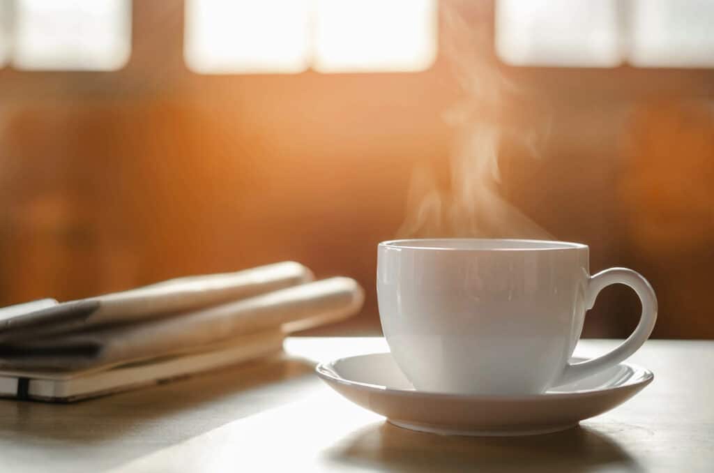 White coffee cup and saucer with steam rising from the cup with newspaper on the table, windows in soft focus in the background.