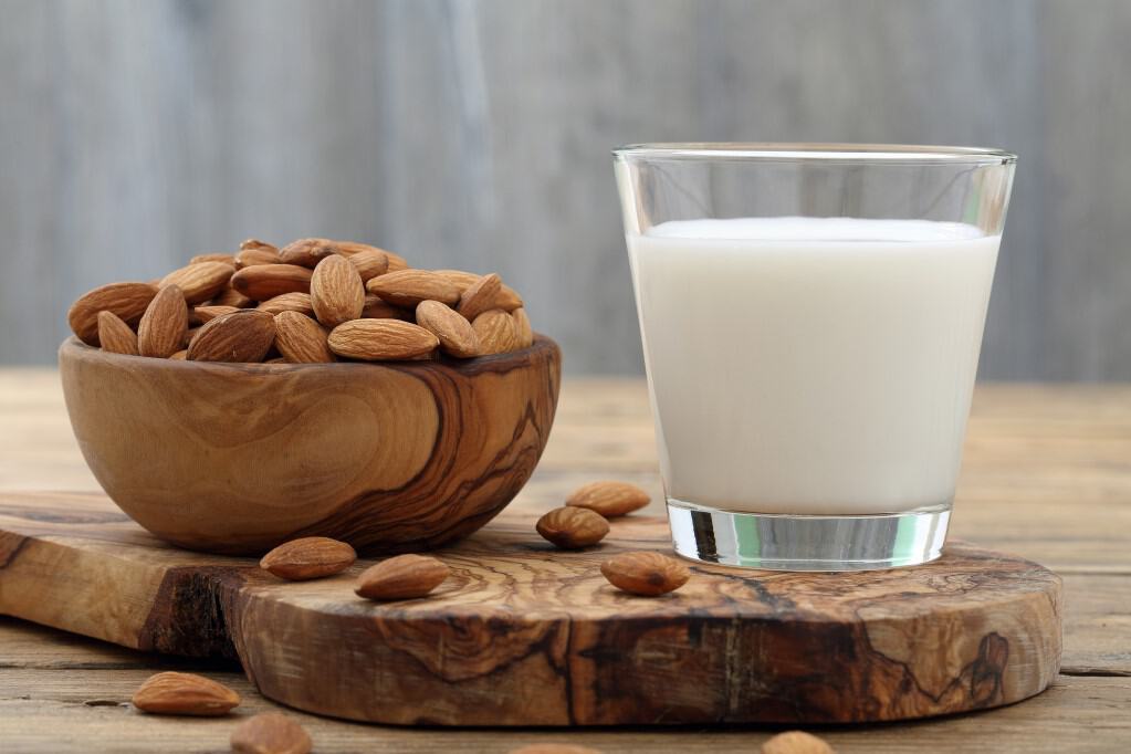 Front view of a small clear glass of almond milk, a natural bowl overflowing with almonds onto a natural wood grain cutting board, all resting on a rustic work surface.