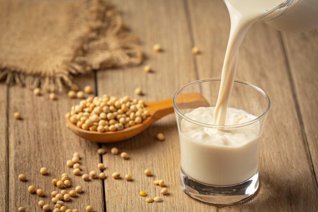 Diagonal view of soy milk pouring from a glass pitcher into a small drinking glass with a medium wooden spoon overflowing with soybeans onto a rustic wooden surface with a folded burlap towel in soft focus in the background.
