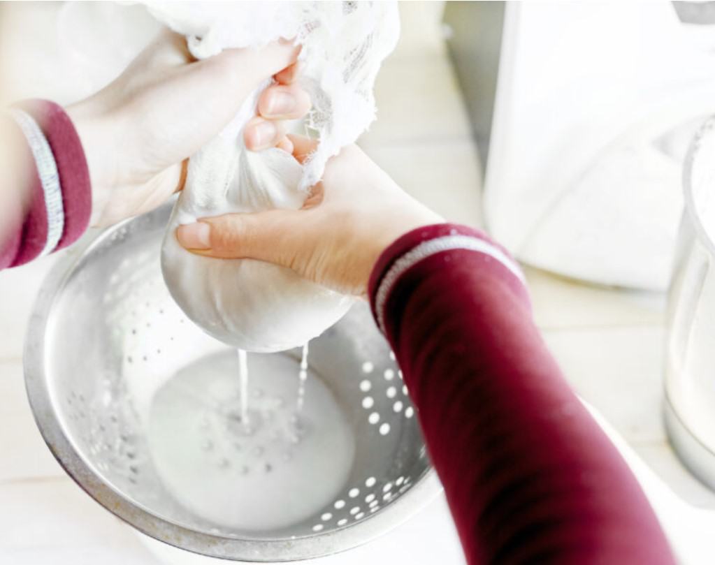 Aerial diagonal view of a Caucasian woman's arms and hands squeezing fresh almond milk out of a cheese cloth into a silver metal colander on a whitewashed surface with a glass pitcher in soft focus in the right of the frame.