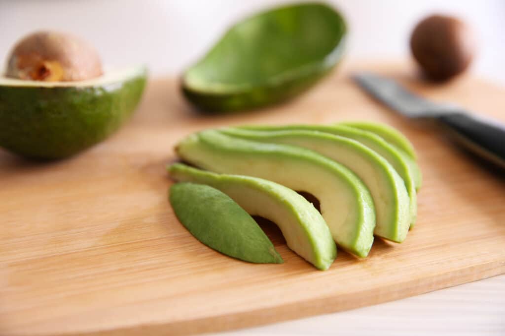 Diagonal view of sliced ripe avocado on a light colored cutting board with a sharp knife, an empty half of avocado skin, an avocado seed and a half of avocado with flesh and seed side up in soft focus in the background, all resting on a light white washed wooden surface. 