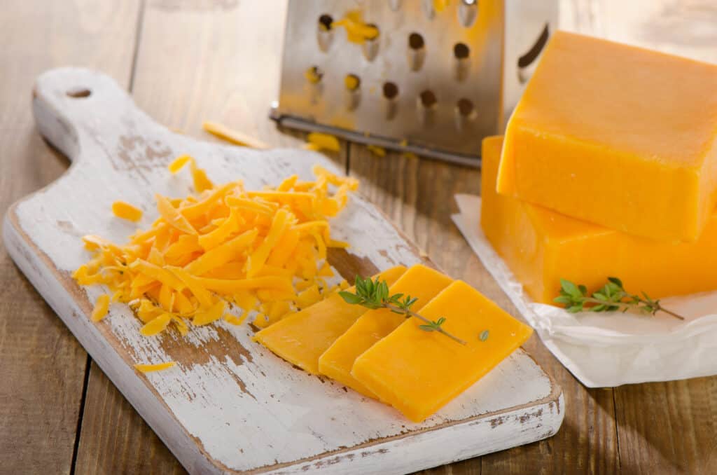 Grated Cheddar Cheese  and 3 slices of cheddar cheese with a spring of fresh herbs on a rustic white wooden Board with two blocks of cheddar cheese in soft focus to the right of the image.
