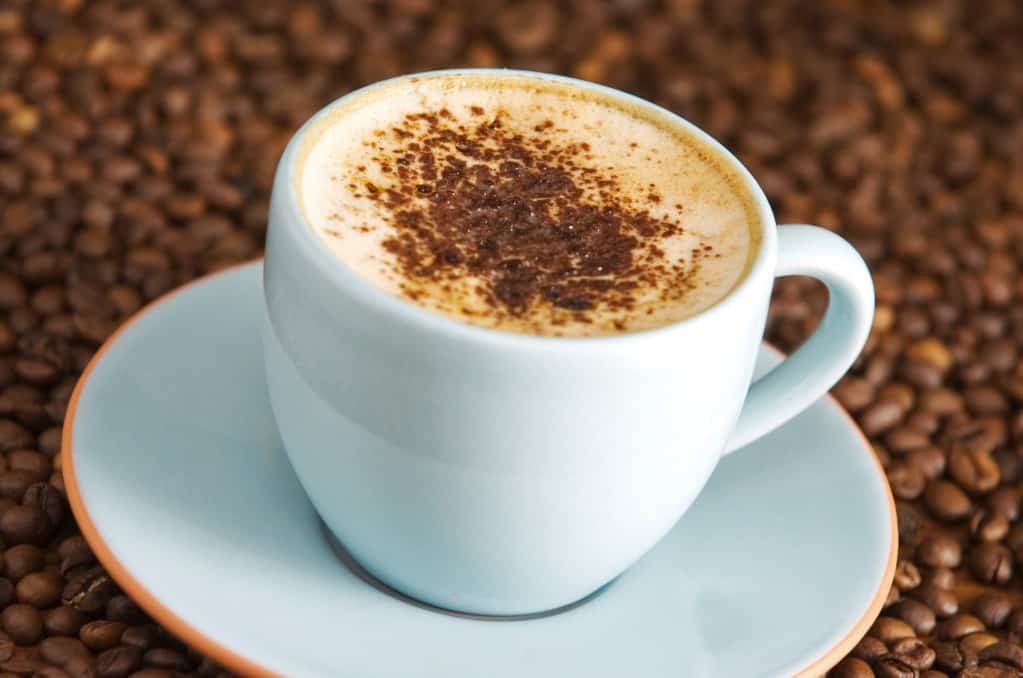 White cappuccino cup and saucer filled with expresso topped with foam milk sprinkled with chocolate powder, with the background and surface filled in with whole coffee beans.