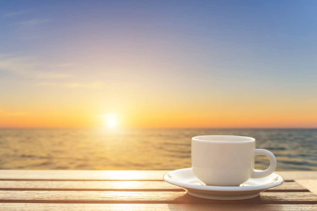 View of a sunrise over the ocean with a wooden table and a white cup and saucer in the foreground.