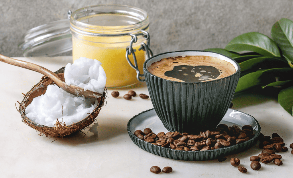 Diagonal view of a dark handle-less cup and saucer filled with coffee and coconut oil with a half coconut shell filled with solid coconut oil and a wooden spoon nearby and a glass jar filled with yellow ghee or butter and a fresh leafy stem of a plant in the background.