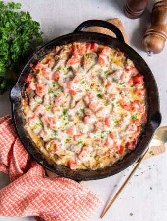 Diagonal view of a close up of Philly cheesesteak casserole in a cast iron pan with a serving being removed by a brass utensil. A red and white kitchen towel is in the foreground and a handful of fresh parsley and wooden salt and pepper grinders are in soft focus in the background.