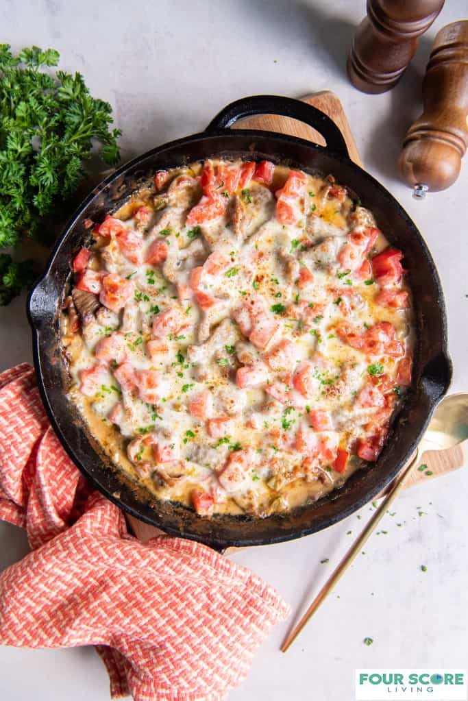 Aerial view of cast iron pan with keto Philly cheesesteak baked inside including sliced steak, cheese and tomatoes, placed on a light wood cutting board with a brass serving utensil next to the cast iron pan and a red and white textured kitchen towel, with a handful of fresh parsley and partial views of wooden salt shaker and pepper grinder, all on a white stone surface.