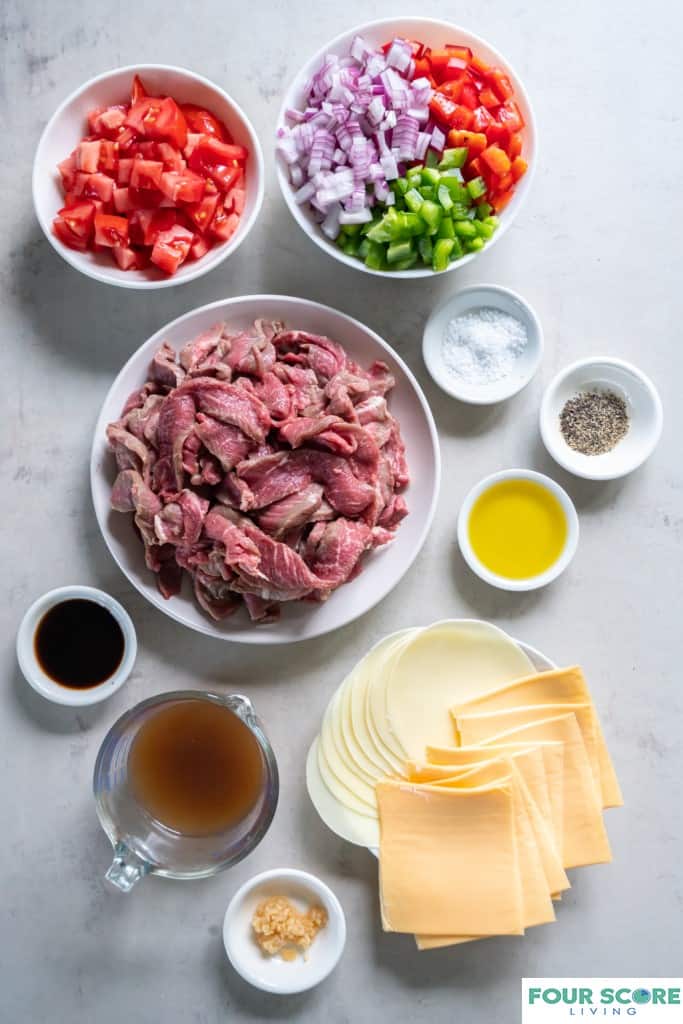Aerial view of ingredients to make Philly cheesesteak casserole set out in different sized white bowls and dishes, including sliced white and yellow cheese, olive oil, minced garlic, tomatoes, sliced raw steak and chopped peppers and onions. 
